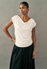 Kermanshahi kaula ka bhawa top jisme chhoti aayen hain, pleating ka design, kale poore skirt ke saath jodi gayi hai. Smooth texture aur aaraamdayak fit. Sunehre cuff bracelet.