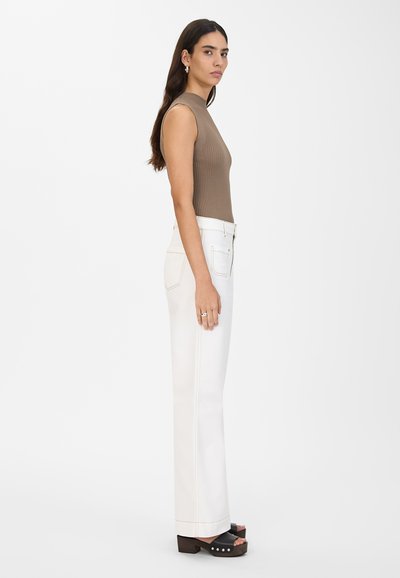 Brun ribbet ermeløs topp kombinert med hvite høytlivsjeans. Modellen har på seg svarte plattformsandaler med hvite detaljer. En ensfarget hvit bakgrunn.
