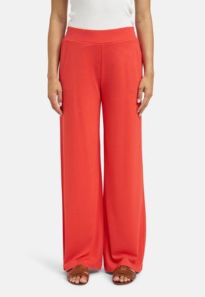 Person wearing high-waisted, wide-leg red pants with a white top and brown open-toe sandals, standing against a plain background.