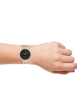 Montre à cadran noir rond avec des index en argent, bracelet en maille rose gold, présentée sur un poignet de peau claire. Design minimaliste, esthétique moderne.