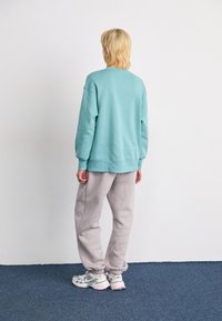 Person med blont hår står vendt mod en hvid væg, iført en teal sweatshirt, lys grå sweatpants og hvide sneakers med pink detaljer.
