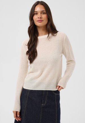 Femme aux longs cheveux foncés portant un pull beige clair et une jupe en jean foncé, debout avec une main dans sa poche.