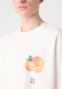 Hvit bomull t-skjorte med et oransje grafisk motiv av en delt appelsin, grønne blader, og "Hugo" tekst i grått under designet.