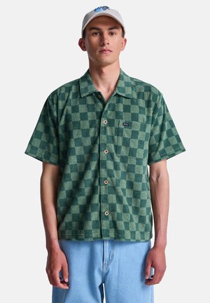 Młody mężczyzna ubrany w zieloną, kraciastą koszulę na krótki rękaw z guzikiem, jasnoniebieskie dżinsy i białą czapkę z niebieskim wzorem.