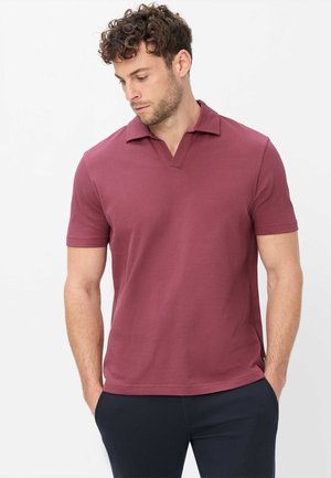 Mann mit lockigem Haar, der ein kurzärmeliges, strukturiertes burgunderfarbenes Poloshirt und dunkle Hosen trägt, die Hände in den Taschen vor einem einfarbigen Hintergrund.