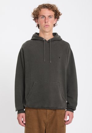 Jeune homme aux cheveux bouclés portant un sweat à capuche gris foncé et un pantalon en velours côtelé marron, debout devant un fond blanc uni.