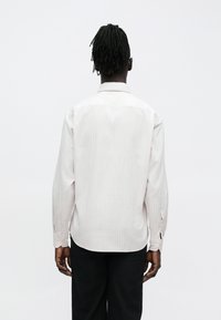 Osoba s tmavými vrkočmi stojí zády, oblečená v svetloružovej pruhovanej blúzke s dlhým rukávom a čiernych nohaviciach na jednoduchom pozadí.