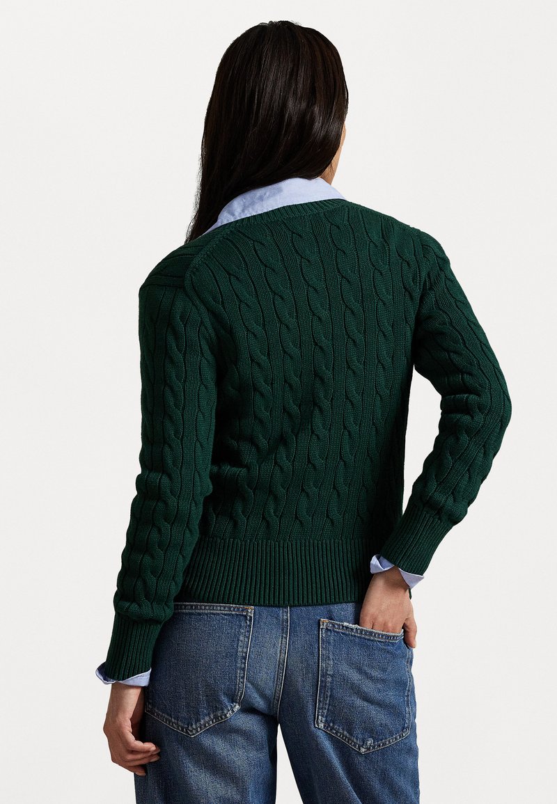 Grøn kabelstrikket sweater med ribbestrikket kant og manchetter, båret over en lyseblå skjorte med krave, set fra bagsiden.