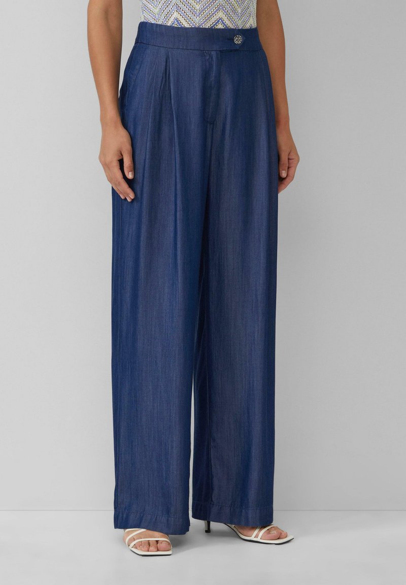 Vrije pijpen donkere denim broek met plooien, een platte tailleband met een metalen knop en een gladde structuur. Draagt met witte sandalen.