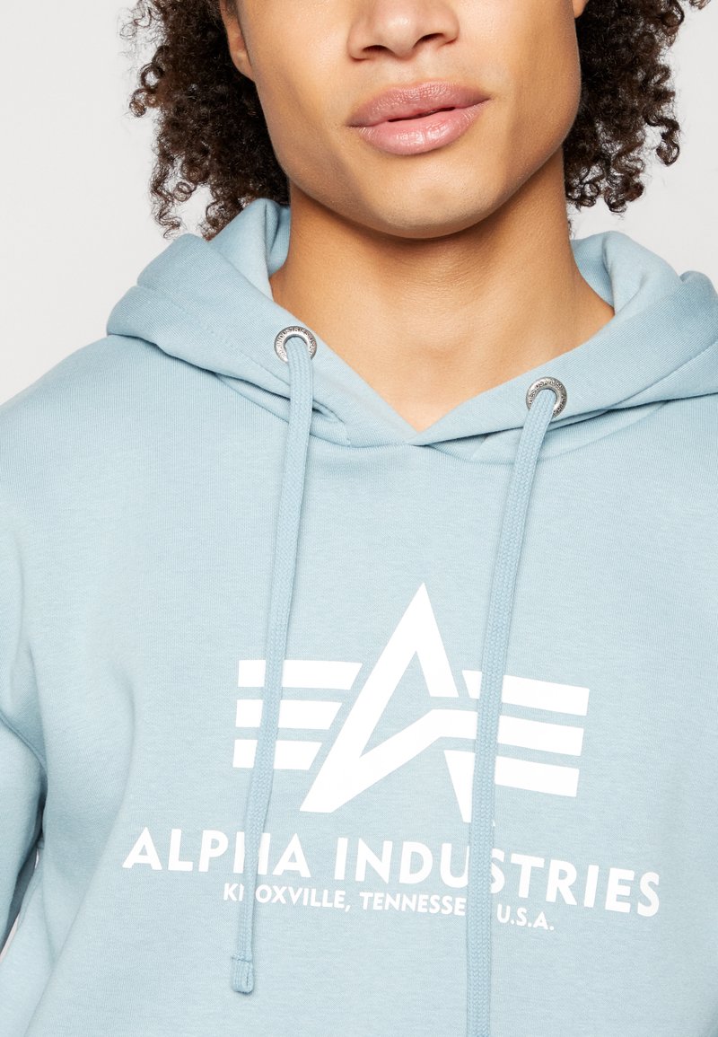 Lichtblauwe hoodie met een trekkoord, voorzien van een wit logo en de tekst "ALPHA INDUSTRIES" prominent weergegeven op de voorkant.