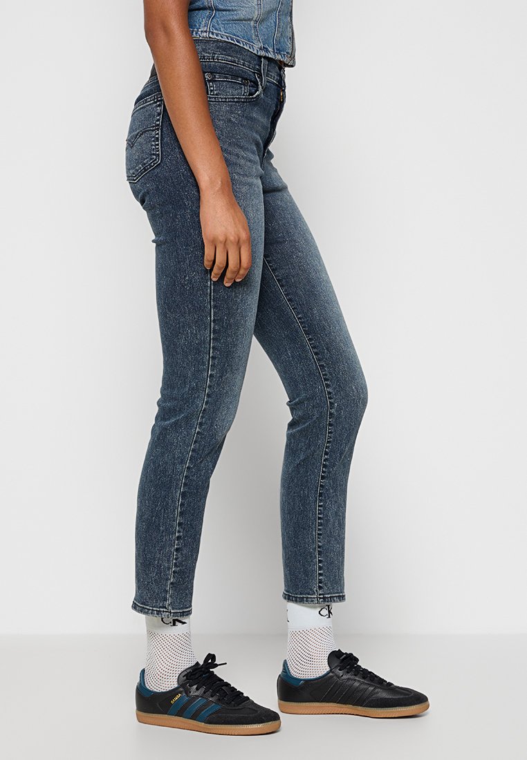 Personne portant un jean slim-fit bleu marine, des chaussettes blanches avec logo et des sneakers Adidas Samba noires avec des semelles en gomme, debout sur un sol blanc uni.