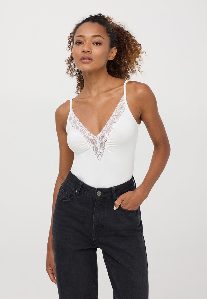 Body bianco con dettaglio in pizzo a V, spalline sottili, abbinato a jeans neri a vita alta. Tessuto liscio con una silhouette aderente.
