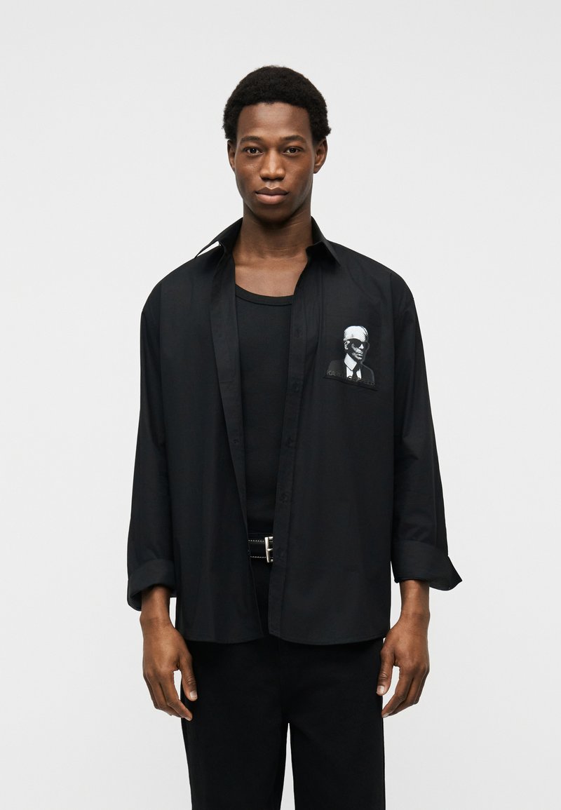 Νεαρός άνδρας που φοράει μια φαρδιά μαύρη πουκάμισο με κουμπιά πάνω από μια μαύρη φανέλα, με ένα μικρό γραφικό ενός άνδρα με γυαλιά ηλίου στην τσέπη της πουκάμισας.