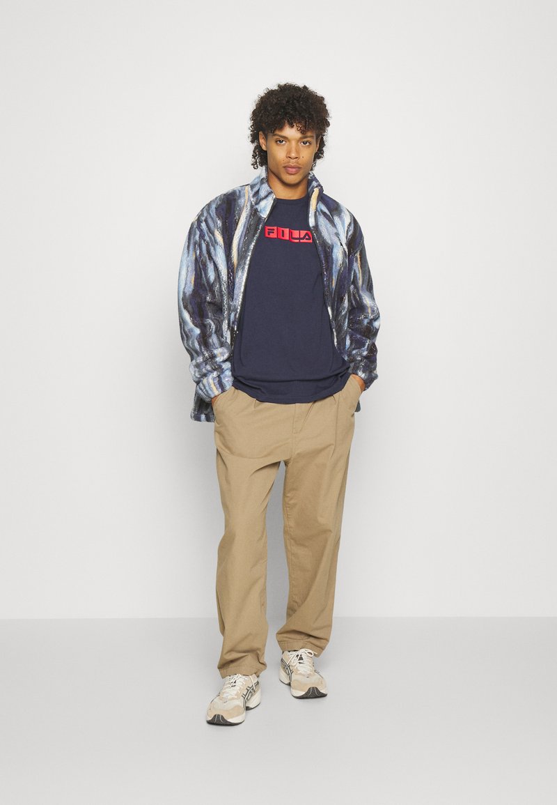 Giacca in pile con motivo a spirale blu e bianco, t-shirt navy con logo rosso "FILA", pantaloni beige e sneakers color crema.