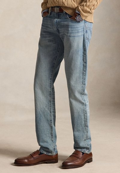 Person iført lyseblå jeans med lige ben, brune læderloafers, brunt bælte, beige sweater og et armbåndsur med brun rem.