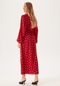 Abito maxi rosso con fiori, maniche lunghe a sbuffo e vita corsetta. Tessuto leggero con motivi di fiori bianchi. Indossato con tacchi neri.