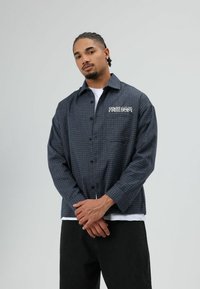 Overhemd met knopen in donkerblauw met een ruitpatroon. Heeft een patch met de tekst "FREEDOM" op de borst en lange mouwen.