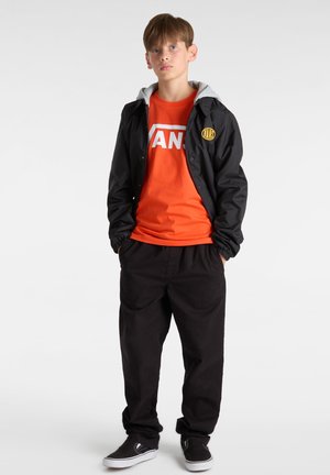 Svart vindjacka med grå huva, orange t-shirt med vit "VANS" logga, svarta byxor och svarta slip-on skor. Avslappnad outfit.