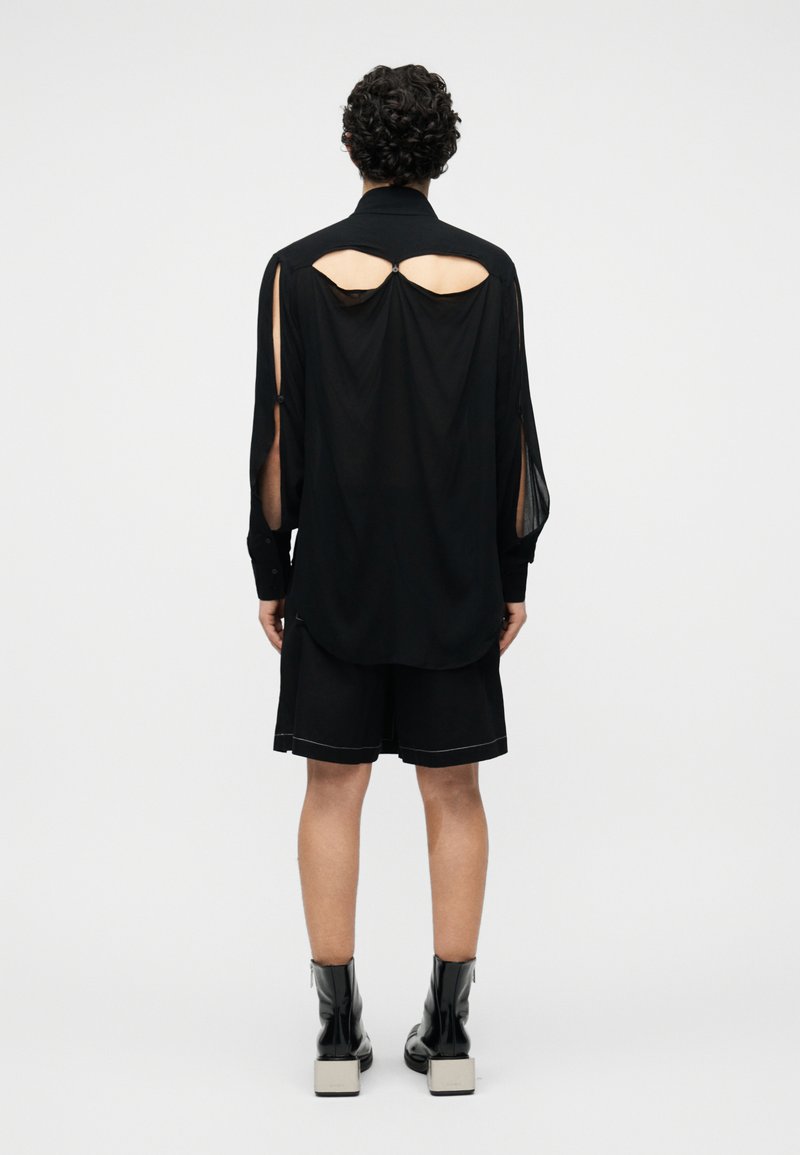 Person with curly hair wears a black long-sleeve top with back cutouts, black shorts, and black ankle boots with white block heels, standing facing away.