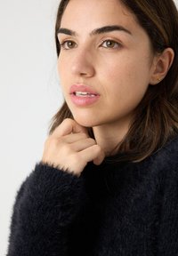 Suéter negro y peludo con un escote alto. Primer plano de una persona descansando la barbilla en la mano. El tono de piel es claro; los labios son de un rosado suave.