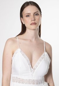 Vestido blanco con tirantes finos, que presenta un escote en V profundo, detalles de encaje y acentos texturizados a lo largo de los bordes. Diseño simple y elegante.