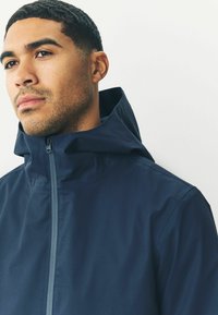 Chaqueta impermeable azul marino con collar alto y cierre con cremallera. Presenta un diseño elegante y una textura suave, adecuada para diversas condiciones climáticas.