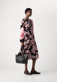 Mujer vistiendo un vestido midi negro con grandes estampados florales en rosa, sosteniendo un bolso de cuero negro y usando zapatos planos negros.