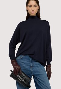 Marinblå polotröja med lös passform och långa ärmar, tillsammans med bruna läderhandskar och en svart texturerad kuvertväska.