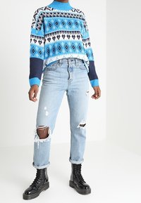 Blauer gemusterter Strickpullover mit hohem Kragen, kombiniert mit hellblauen, zerrissenen Jeans und schwarzen Schnürkampfboots.