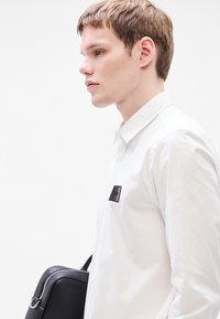 Λευκό πουκάμισο με εφαρμοστή γραμμή και τσέπη με μαύρη ετικέτα, σε συνδυασμό με μια μαύρη δερμάτινη τσάντα με ασημένιο φερμουάρ.