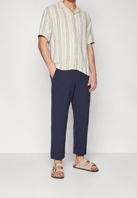 Lyhythihainen, beige-siniraidallinen paita, yhdistetty tummansinisten housujen ja kahdella soljella varustettujen beigen sandaalien kanssa. Rento ilme, väljä istuvuus.