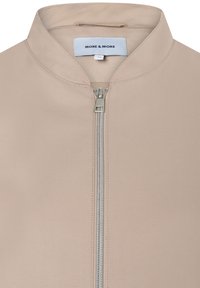 Beige jacket with a high collar, front zipper closure, and smooth fabric texture. Logo tag "MORE & MORE" on the neck. Simple design.