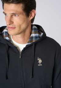 Felpa con zip di colore blu navy, dotata di stringhe, con foderatura in plaid blu, bianco e giallo. Logo ricamato sul petto. Materiale in cotone morbido.