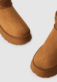 Stivali in suede marrone per l'inverno con suole in gomma spessa e testurizzata, mostrati da un'angolazione superiore-frontale su uno sfondo grigio chiaro.