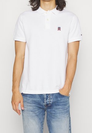 Camisa polo blanca de algodón, con una tapeta de dos botones, mangas cortas y un pequeño logo en el pecho. Se lleva con jeans azules.