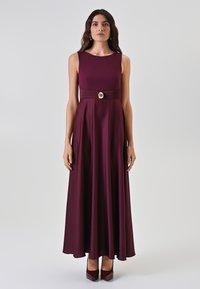 Robe maxi sans manches bordeaux avec un corsage ajusté, une taille plissée et un accent circulaire doré. Tissu lisse avec une jupe fluide.