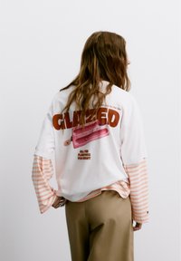Weißes T-Shirt mit einem retro-inspirierten Grafikwurf, das rosa Eis am Stiel und den Text "GLAZED" in fettem Rot zeigt. Lange gestreifte Ärmel blitzen hervor.