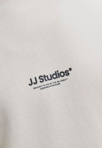 Kremkrāsas audumam ir teksturēta virsma ar tumši zilu logotipu "JJ Studios" un papildu tekstu zem tā.