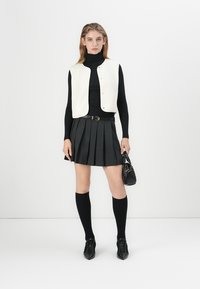 Weißer, ärmelloser Weste über einem schwarzen Rollkragenpullover, kombiniert mit einem schwarzen Faltenrock. Accessoires sind eine kleine schwarze Handtasche und kniehohe Socken.