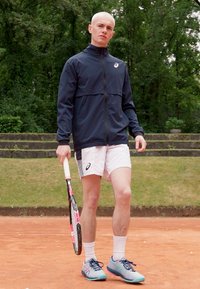 Homme athlétique, portant une veste à fermeture éclair noire, un short blanc et des chaussures de tennis bleu clair, tenant une raquette de tennis avec un accent rose.