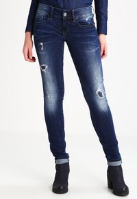 Jeans ajustados en denim azul oscuro con un lavado desgastado. Presenta parches desgastados en las rodillas y dobladillos con vuelta. Cierre de botón metálico y bolsillos delanteros.