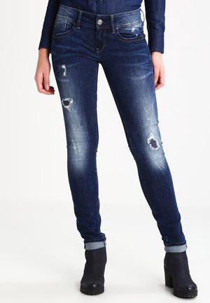 Jeans ajustados en denim azul oscuro con un lavado desgastado. Presenta parches desgastados en las rodillas y dobladillos con vuelta. Cierre de botón metálico y bolsillos delanteros.