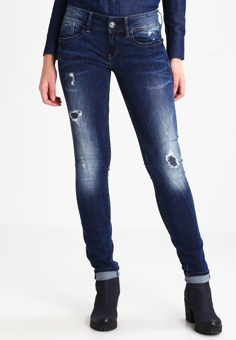 Jeans ajustados en denim azul oscuro con un lavado desgastado. Presenta parches desgastados en las rodillas y dobladillos con vuelta. Cierre de botón metálico y bolsillos delanteros.
