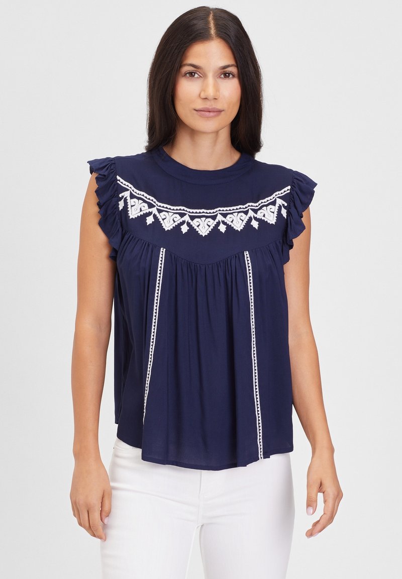 Marineblaues, kurzärmliges Oberteil mit Rüschenschultern, das weiße gestickte Muster und ein plissiertes Design aufweist. Kombiniert mit weißen Hosen.