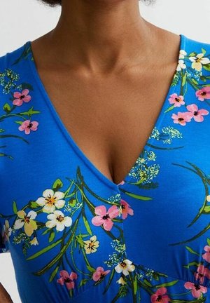 Femme portant une robe bleue à col en V avec des motifs floraux roses, blancs et jaunes et des accents feuillus verts.