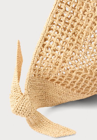 Κοντινό πλάνο μπεζ πλεκτού ψάθινου καπέλου με υφή πλεκτού μοτίβου και λεπτομέρεια με δεμένο φιόγκο σε ουδέτερο φόντο.