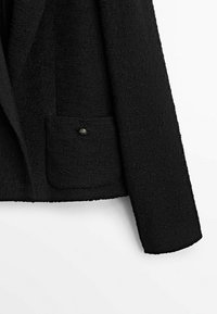 Musta tekstuuriltaan blazer, jossa on yksi etutasku koristeellisin nappi. Kangas on tiheä ja hieman koholla, mikä lisää sen visuaalista kiinnostavuutta.