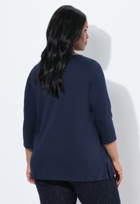 Navyblauwe top met driekwartmouwen, met zijsplitten en een soepele textuur. Getoond vanaf de achterkant tegen een witte achtergrond.