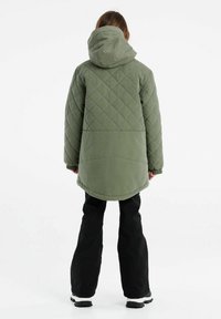 Giacca trapuntata verde con cappuccio, caratterizzata da un orlo posteriore più lungo, polsini elasticizzati, abbinata a pantaloni neri e stivali.