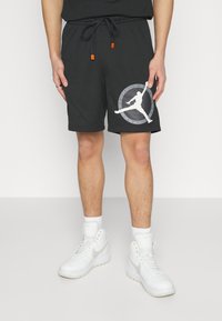 Shorts de basketball noirs avec un motif texturé, présentant un grand logo blanc sur la jambe gauche et des accents de cordon orange.